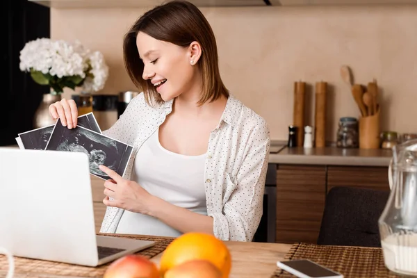 Selective focus of happy pregnant woman pointing with finger at ultrasound photos while having video call — Stock Photo