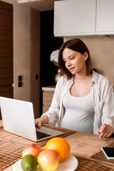 Selective focus of pregnant freelancer using laptop near fruits on table — Stock Photo