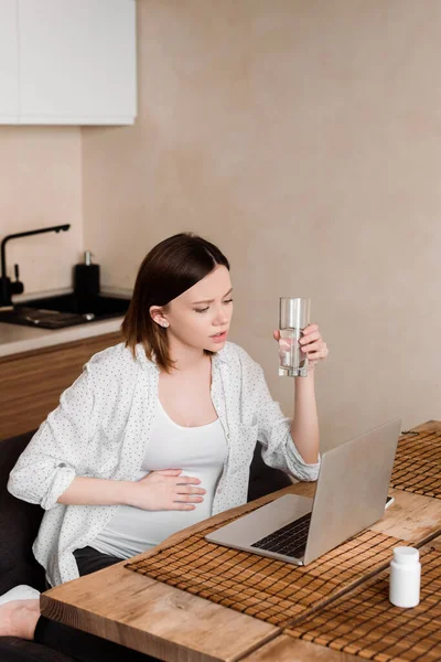 Freelancer embarazada sosteniendo vaso de agua y tocando el vientre cerca de la computadora portátil — Stock Photo