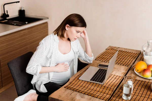 Freelance incinta che tocca la pancia vicino a computer portatile, smartphone, vetro d'acqua e frutta sul tavolo — Foto stock
