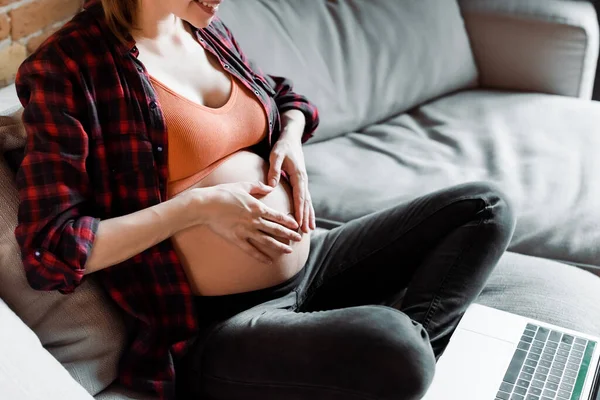 Vista cortada da mulher grávida tocando barriga e sorrindo ao ter chamada de vídeo na sala de estar — Fotografia de Stock