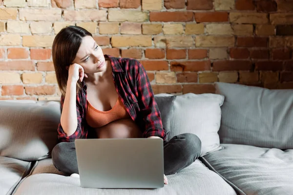 Disgustada y embarazada freelancer mirando hacia otro lado cerca del ordenador portátil en la sala de estar - foto de stock