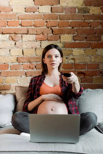 Mujer embarazada sosteniendo el mando a distancia y viendo la película cerca del ordenador portátil - foto de stock