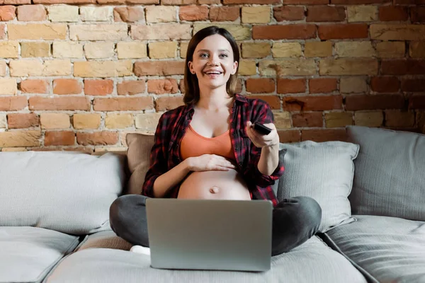 Happy and pregnant woman holding remote controller while watching movie near laptop — Stock Photo