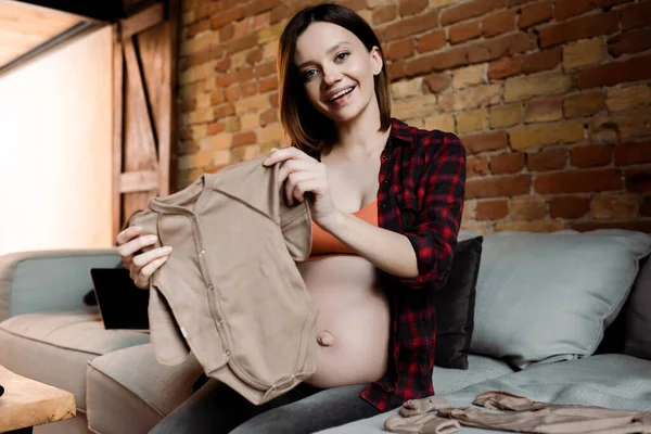 Cheerful and pregnant woman holding baby romper and sitting on sofa — Stock Photo