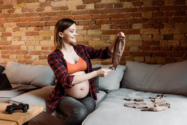 KYIV, UKRAINE - 30 AVRIL 2020 : femme gaie et enceinte tenant un pantalon bébé près d'un joystick sur la table — Photo de stock