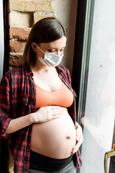 Femme enceinte en masque médical toucher le ventre près de la fenêtre — Photo de stock