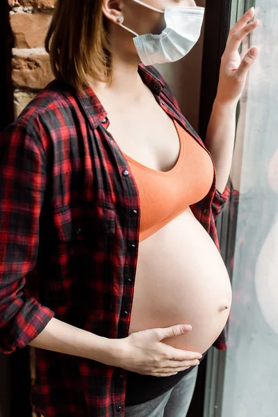 Vista recortada de la mujer embarazada en máscara médica tocando el vientre cerca de la ventana - foto de stock
