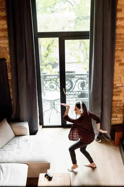 KYIV, UCRANIA - 30 DE ABRIL DE 2020: Mujer alegre y embarazada en auriculares inalámbricos bailando y escuchando música en la sala de estar - foto de stock