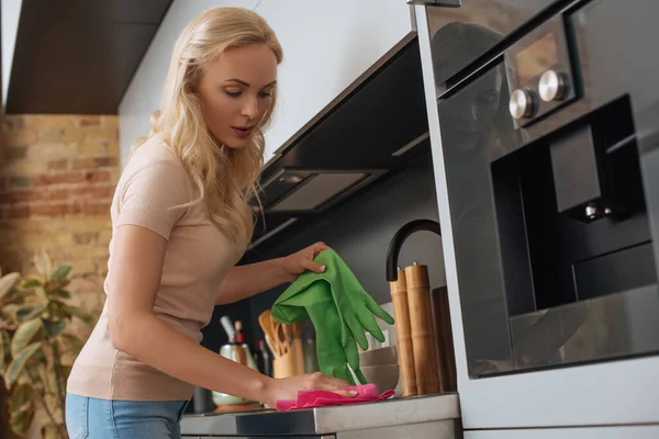 Dona de casa atraente segurando luvas de borracha enquanto limpa a superfície da cozinha com pano — Fotografia de Stock