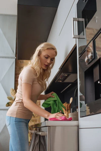 Joven ama de casa sosteniendo guantes de goma mientras limpia la superficie de la cocina con trapo - foto de stock