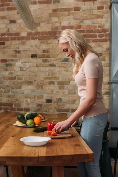 Vue latérale de la jeune femme blonde coupant des légumes frais dans la cuisine — Photo de stock