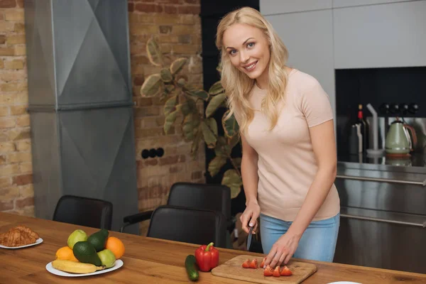 Mujer feliz mirando a la cámara mientras corta verduras frescas en la tabla de cortar - foto de stock