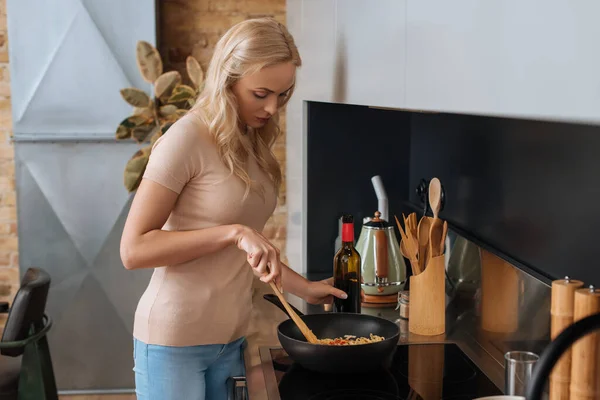 Junge blonde Frau hält eine Flasche Rotwein in der Hand, während sie Thai-Nudeln im Wok zubereitet — Stockfoto