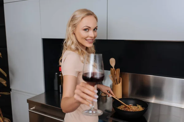 Selektiver Fokus einer jungen Frau mit einem Glas Rotwein und einem Lächeln in die Kamera, während sie thailändische Nudeln zubereitet — Stockfoto