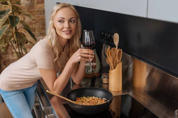 Sonriente mujer con copa de vino tinto de pie cerca de wok con fideos tailandeses y mirando hacia otro lado - foto de stock