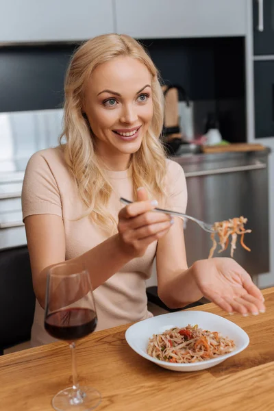 Mujer sonriente sosteniendo tenedor con fideos tailandeses cerca de un vaso de vino tinto - foto de stock