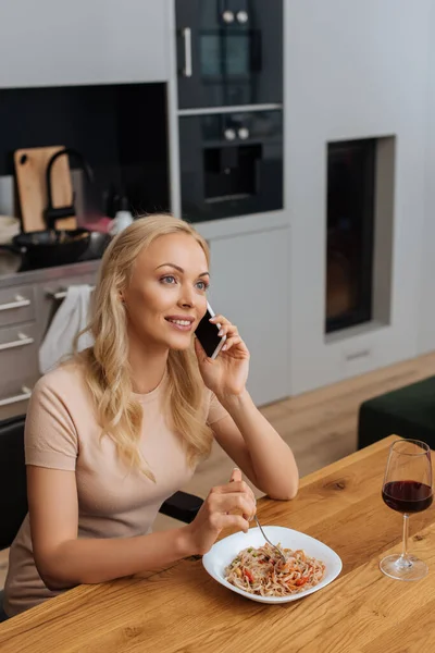 Mujer sonriente hablando en el teléfono inteligente mientras se sienta cerca del plato con fideos tailandeses y una copa de vino tinto - foto de stock