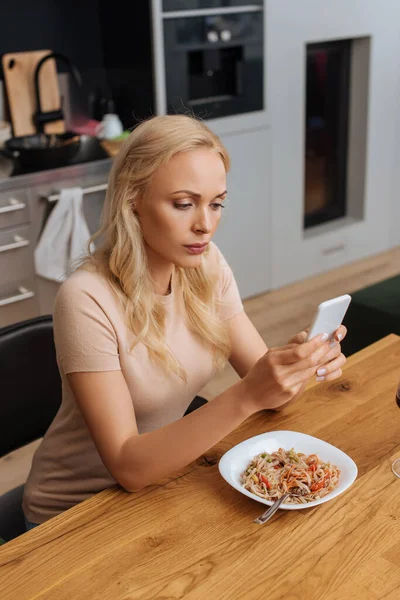 Ernste Frau plaudert auf Smartphone, während sie neben Teller mit Thai-Nudeln sitzt — Stockfoto