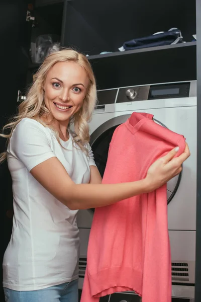 Ama de casa sonriente mirando a la cámara mientras sostiene el jersey cerca de la lavadora - foto de stock