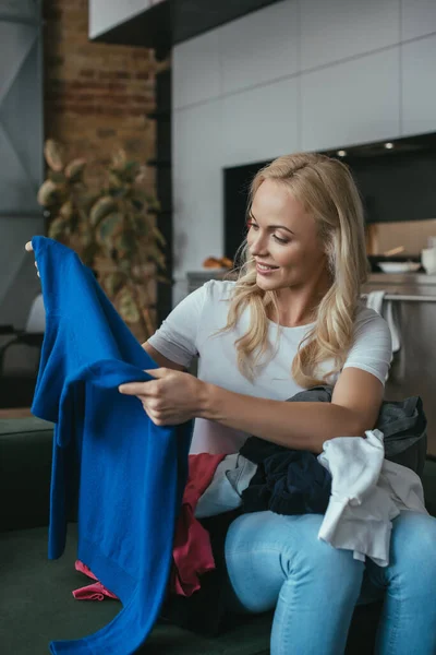 Lächelnde Hausfrau blickt auf Pullover, während sie mit Wäsche auf dem Schoß sitzt — Stockfoto