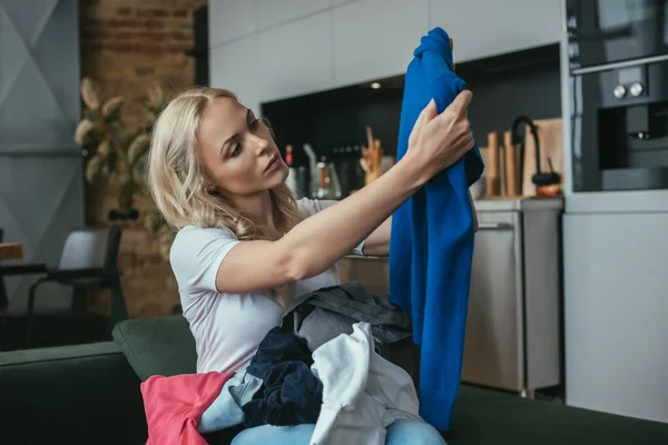 Junge Hausfrau schaut Pullover an, während sie mit Wäsche auf dem Schoß sitzt — Stockfoto