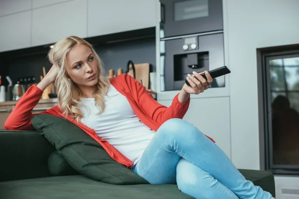 Mulher séria sentado no sofá e segurando tv controlador remoto — Fotografia de Stock