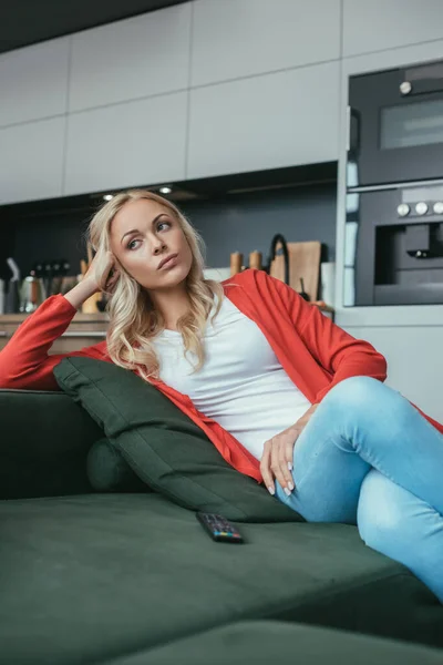Nachdenkliche Frau schaut weg, während sie auf dem Sofa in der Nähe der Fernbedienung sitzt — Stockfoto