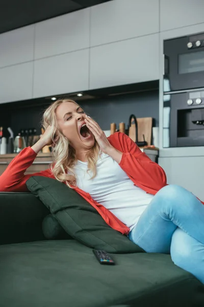 Mulher sonolenta cobrindo a boca com a mão enquanto bocejando no sofá perto de tv controlador remoto — Fotografia de Stock
