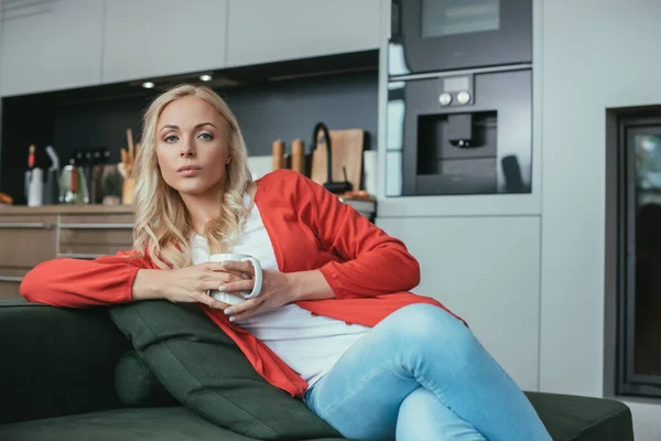 Mujer seria mirando a la cámara mientras está sentado en el sofá con una taza de té — Stock Photo