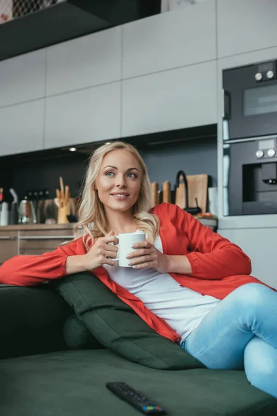 Donna felice che distoglie lo sguardo mentre siede sul divano con una tazza di tè — Foto stock