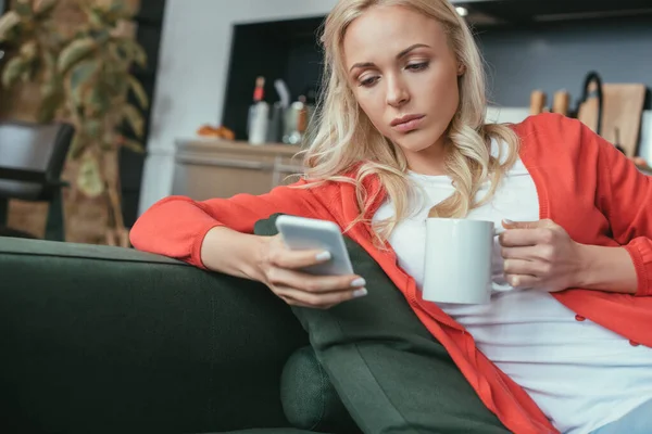Femme réfléchie bavarder sur smartphone tout en étant assis sur le canapé avec une tasse de thé — Photo de stock