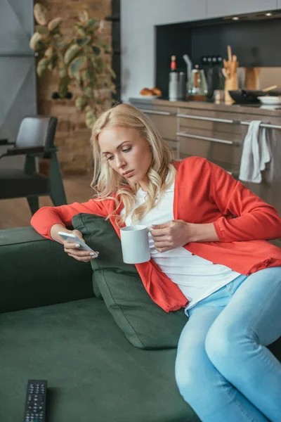 Serious woman chatting on smartphone while sitting on sofa with cup of tea — Stock Photo