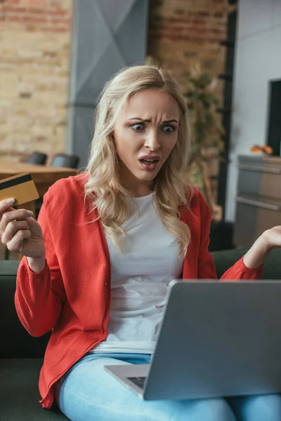 Mujer sorprendida mirando a la computadora portátil mientras sostiene la tarjeta de crédito - foto de stock