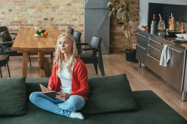 Vue grand angle de femme rêveuse assise sur le canapé, tenant le livre et regardant loin — Photo de stock