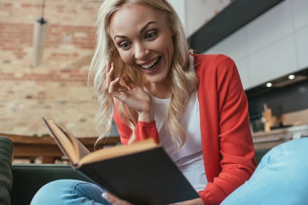 Selective focus of excited woman laughing while reading book at home — Stock Photo