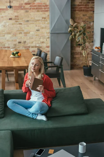 Vue grand angle de femme coûteuse assise sur le canapé, tenant le livre et regardant loin — Photo de stock