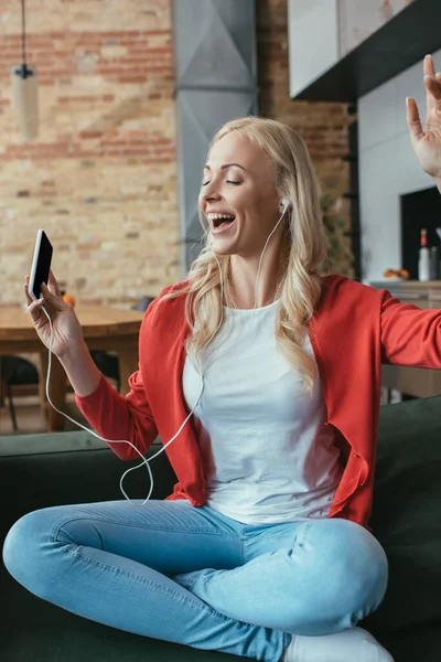 Felice donna in auricolare cantare ad occhi chiusi e alzata di mano mentre tiene lo smartphone — Foto stock