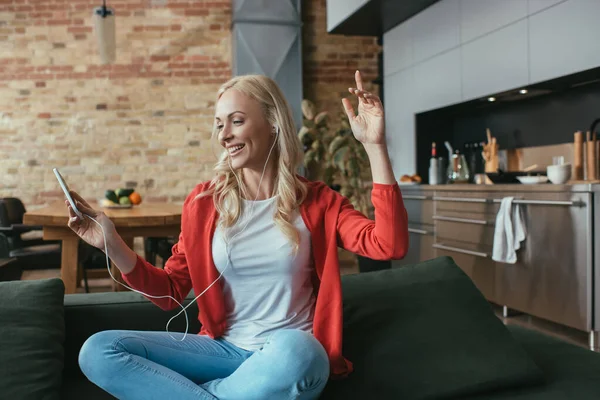 Cheerful woman listening music in earphones while sitting with raised hand and holding smartphone — Stock Photo