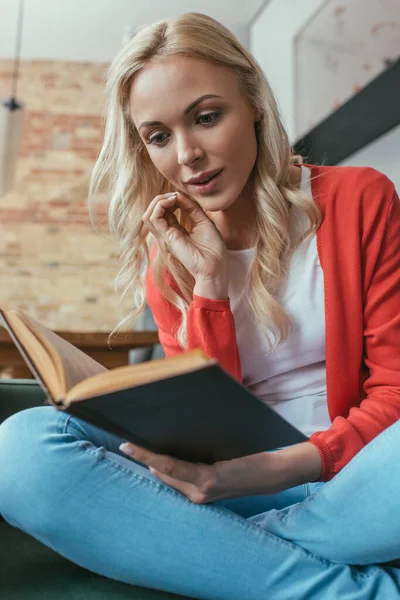 Aufmerksame blonde Frau hält die Hand vor das Gesicht, während sie zu Hause Buch liest — Stockfoto