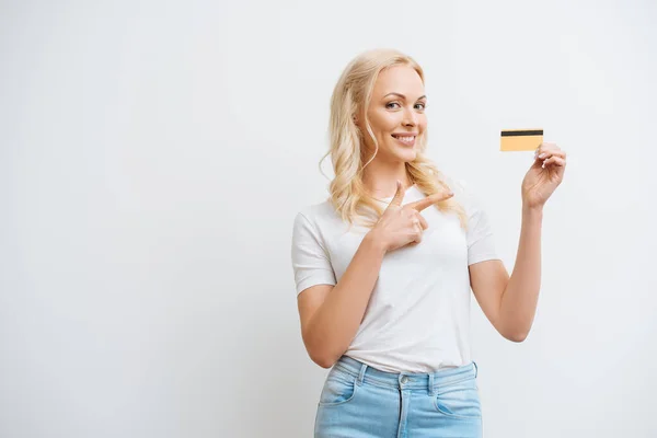 Mulher alegre apontando com o dedo para o cartão de crédito enquanto olha para a câmera isolada no branco — Fotografia de Stock