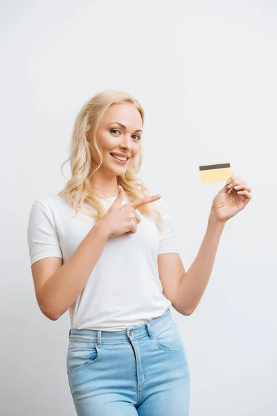Happy woman pointing with finger at credit card while looking at camera isolated on white — Stock Photo