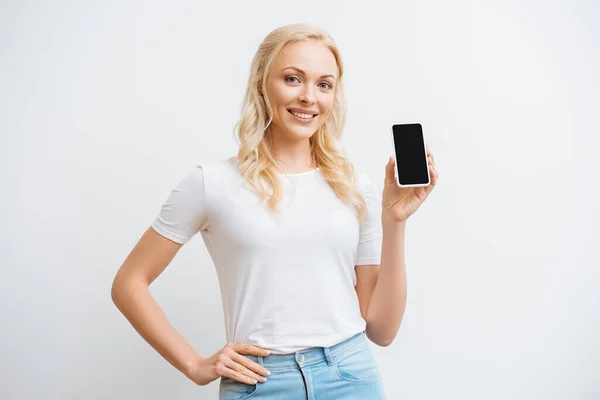 Mujer feliz cogida de la mano en la cadera mientras muestra el teléfono inteligente con la pantalla en blanco y sonriendo a la cámara aislada en blanco — Stock Photo