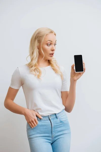Здивована жінка тримає руку в кишені, дивлячись на смартфон з порожнім екраном ізольовано на білому — стокове фото