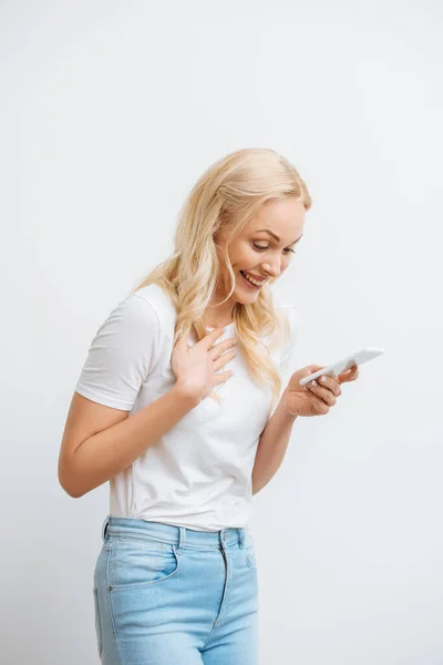 Aufgeregtes Mädchen hält Hand auf Brust und lacht während Videochat auf Smartphone isoliert auf weiß — Stockfoto