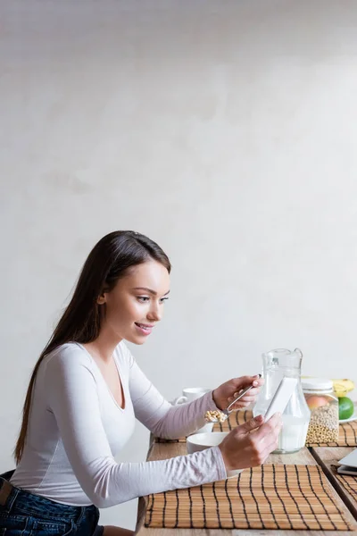 Menina feliz usando smartphone e colher segurando com saborosos flocos de milho — Fotografia de Stock