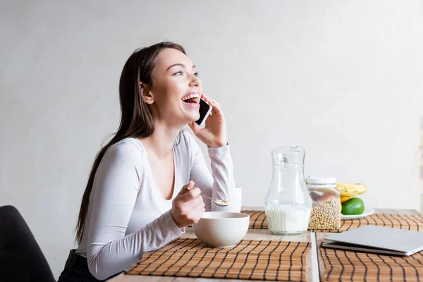 Chica feliz hablando en el teléfono inteligente y riendo mientras sostiene la cuchara con sabrosos copos de maíz - foto de stock