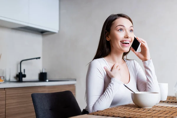 Alegre chica hablando en el teléfono inteligente y riendo cerca de sabrosos copos de maíz en tazón - foto de stock