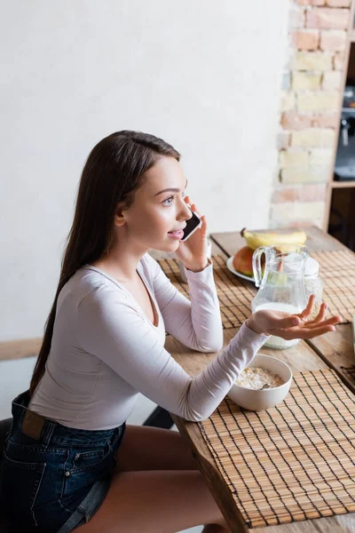 Chica atractiva hablando en el teléfono inteligente y el gesto cerca de sabrosos copos de maíz en un tazón - foto de stock
