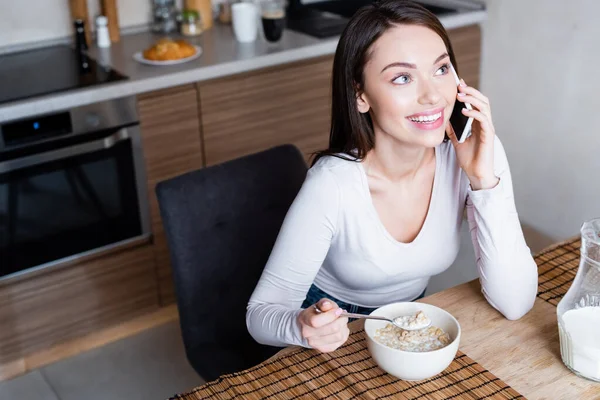 Chica feliz hablando en el teléfono inteligente y riendo mientras sostiene la cuchara con deliciosos copos de maíz y leche - foto de stock